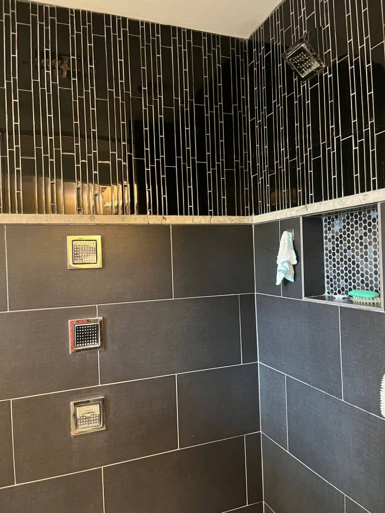 Modern shower interior with dark rectangular wall tiles, three square shower jets, a built-in niche holding a washcloth, and a rainfall showerhead on the ceiling—an award-winning remodel designed for luxury and comfort.