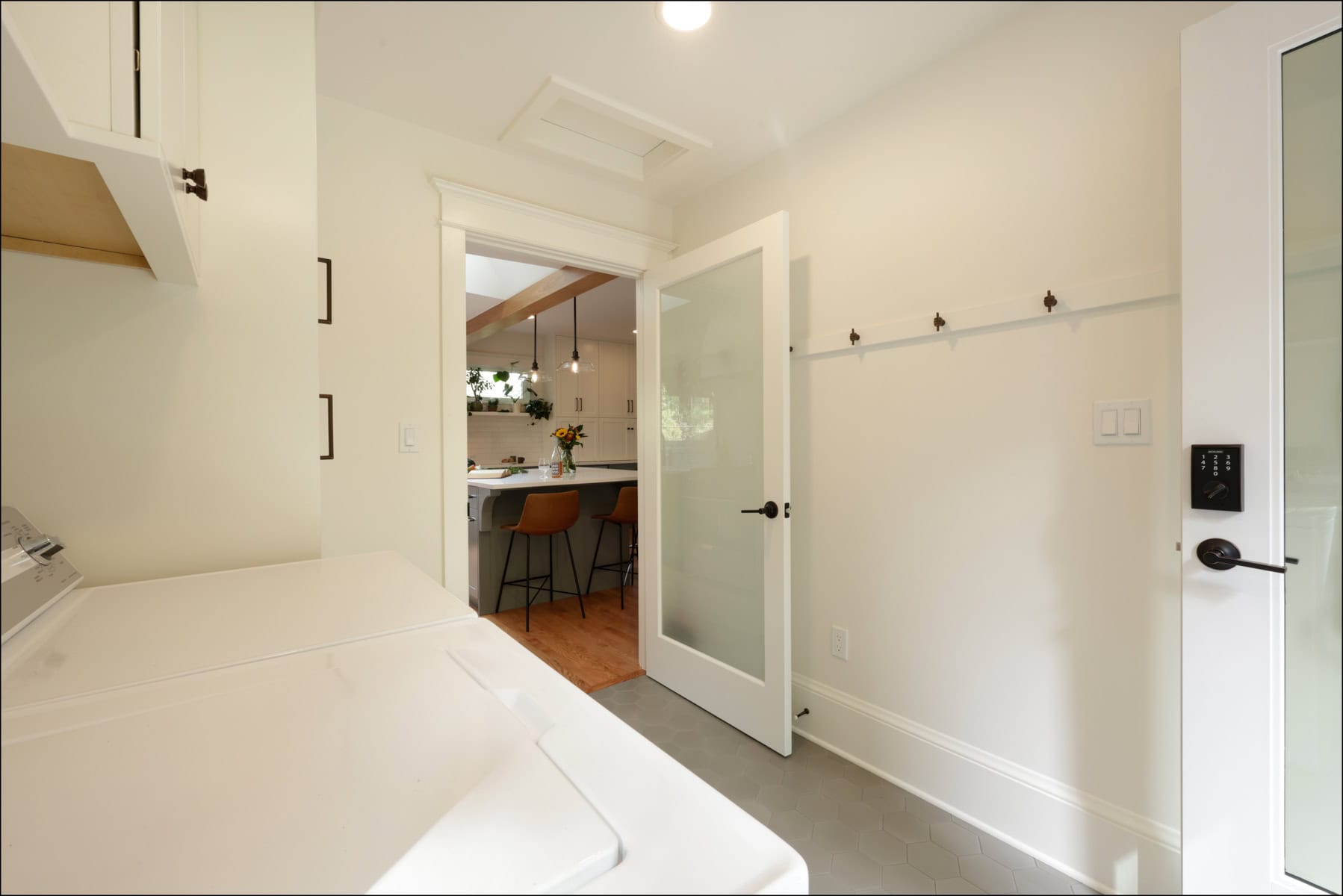 A laundry room with white appliances and a glass door opens to a kitchen with bar stools, wooden floor, and visible ceiling beams.