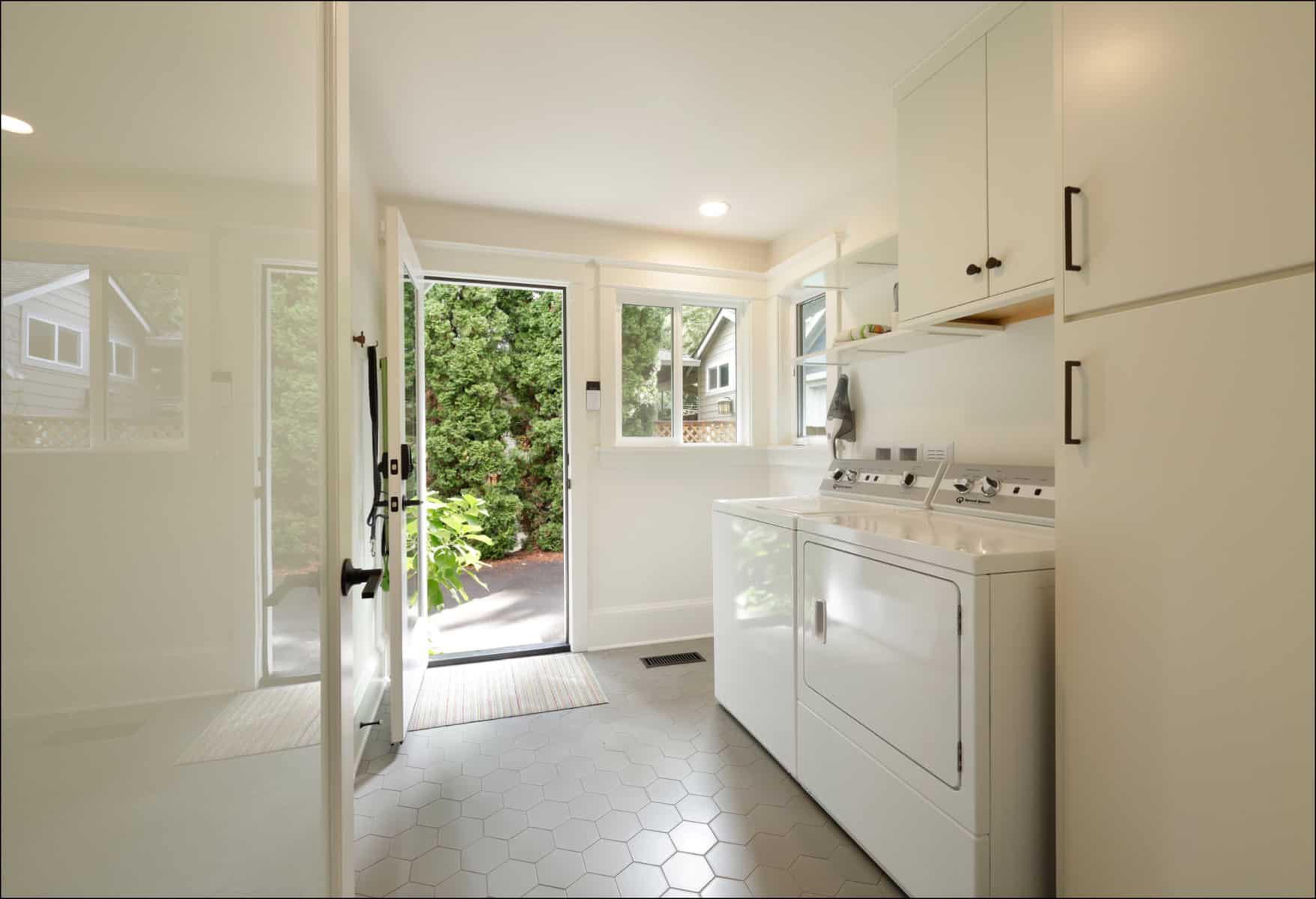 Bright laundry room with white cabinets, a washer and dryer, hexagonal floor tiles, and a door open to a garden outside.