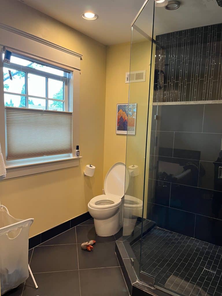 A bathroom with a toilet, a glass shower, window with blinds, tiled floor and walls, laundry basket, toilet paper roll, and a framed picture—crafted by an award-winning remodeler for exceptional style and comfort.