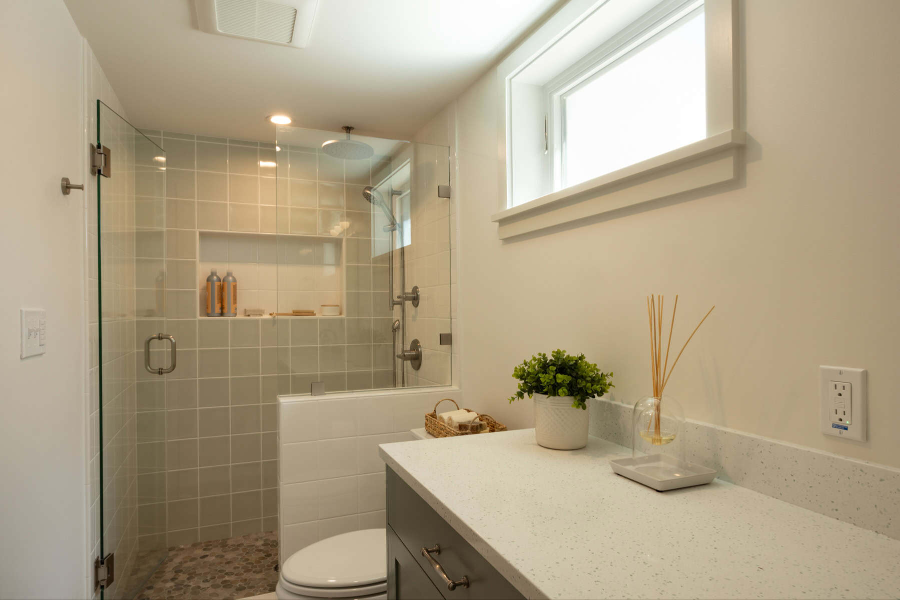 Modern bathroom with a glass-enclosed shower, white tiled walls, a window, and a countertop with a potted plant and reed diffuser.