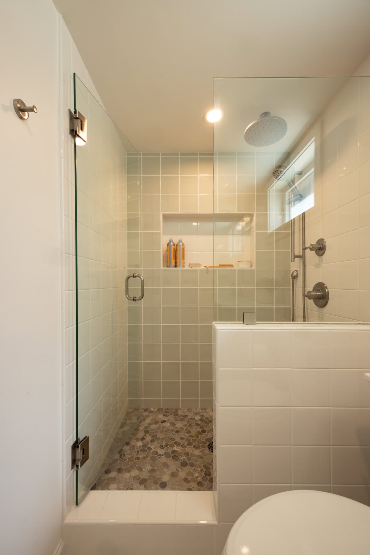 Modern shower with glass door, tiled walls, pebble floor, built-in shelf holding toiletries, and a rainfall showerhead. Partial view of a toilet in the foreground.