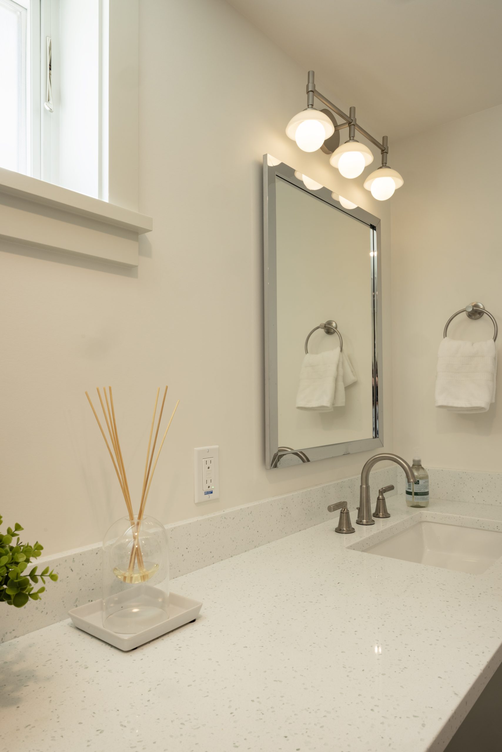 Modern bathroom vanity with a white countertop, a rectangular mirror, three-bulb light fixture, hand towel, reed diffuser, and small potted plant.