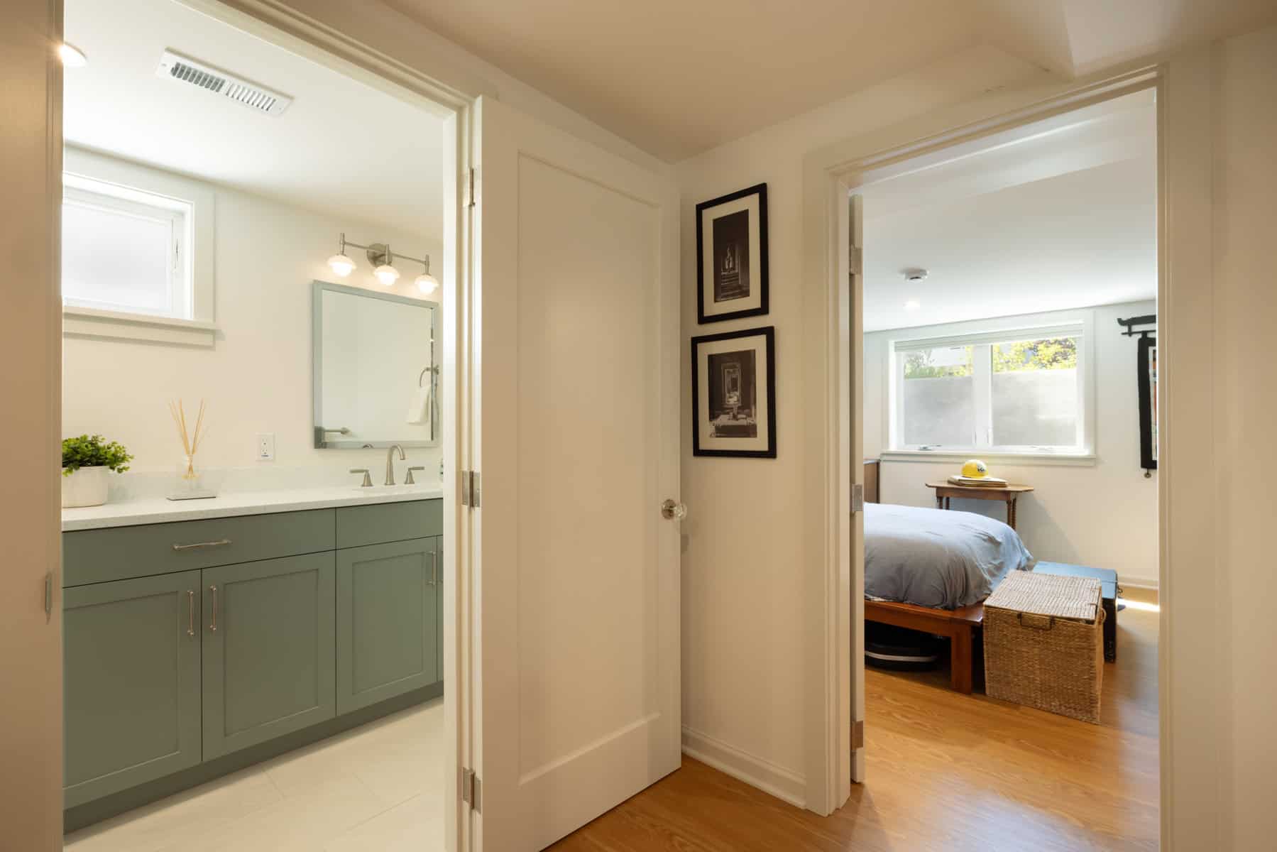 A hallway with doors to a bathroom with a green vanity on the left and a bedroom with wood floors and a wicker chest at the foot of the bed on the right.