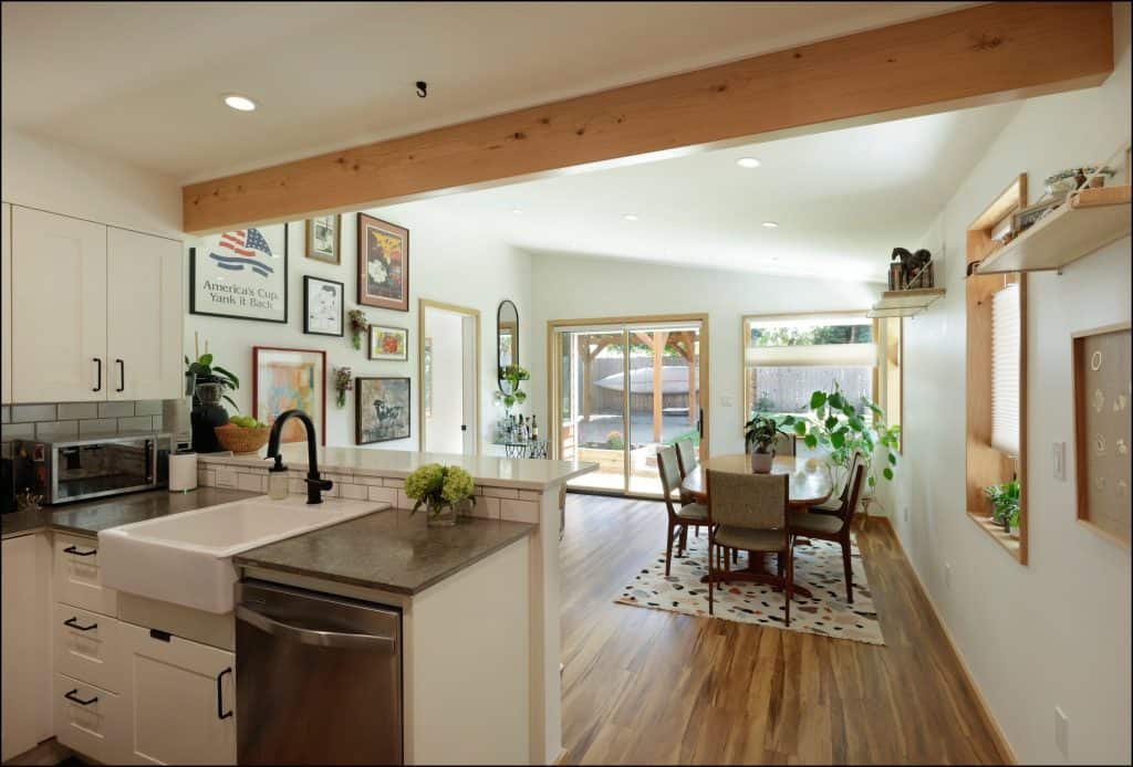 Modern kitchen with a farmhouse sink opens to a dining area featuring wood floors, a round table, and indoor plants. Art decor highlights the importance of design choices in any home remodel, enhanced by large windows and curated shelves.