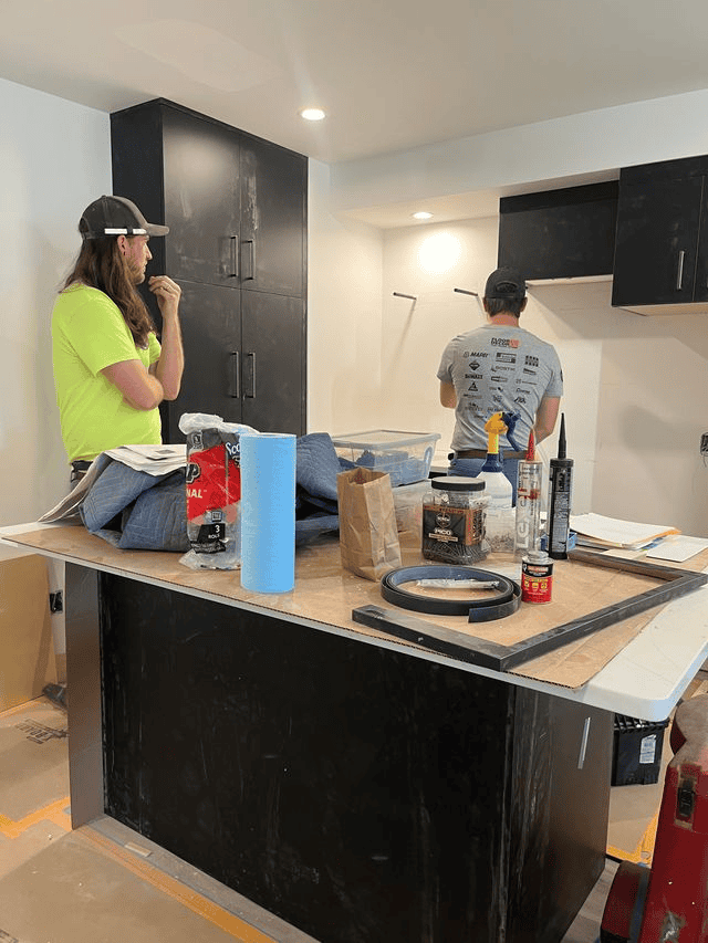 Who are all these people in my house? Two workers, crucial subcontractors in this home remodel, are installing cabinets in a kitchen under renovation; various tools and supplies are spread out on the island countertop.