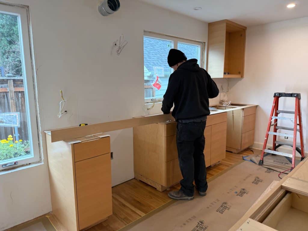 A person in dark clothing installs wooden cabinets and a countertop in a kitchen under renovation, highlighting the importance of skilled subcontractors in a successful home remodel, with tools and a red ladder nearby.