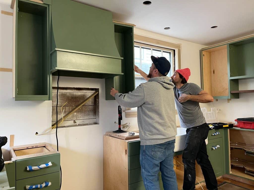 Two people are installing green kitchen cabinets in a partially finished kitchen, highlighting the importance of teamwork during a home remodel, with tools and materials visible around them.