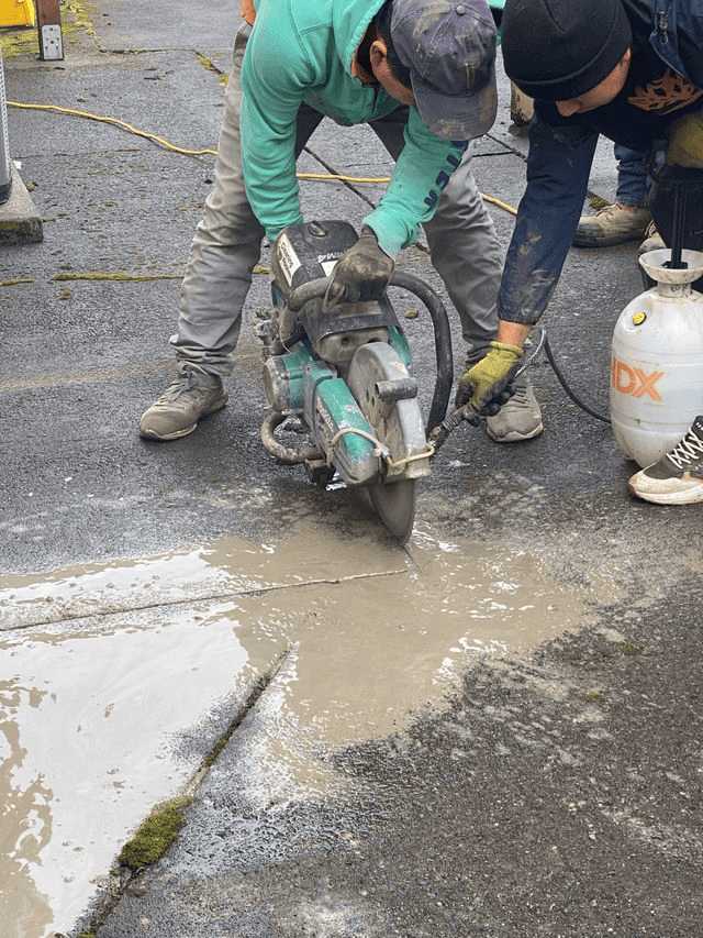 Two subcontractors use a power saw to cut through wet concrete on a paved surface, highlighting the importance of precision and safety as water and debris scatter around the cutting area.