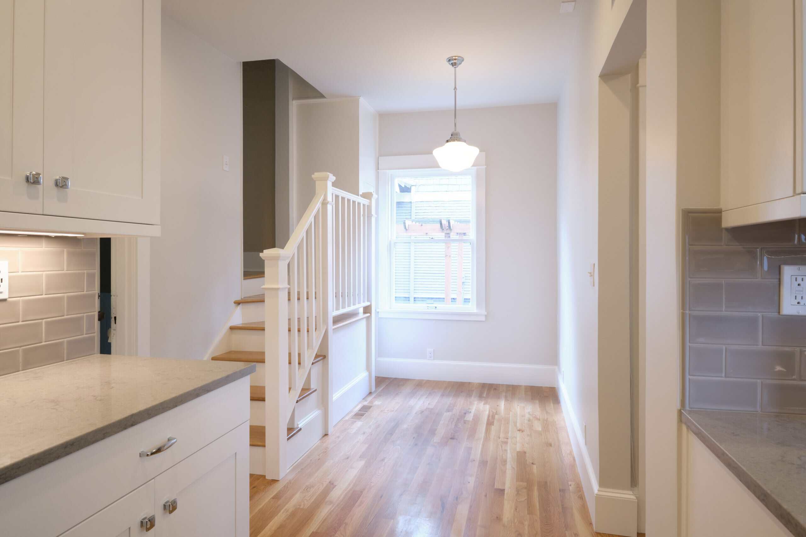 A bright, modern kitchen and dining area with light wood floors, white cabinets, gray countertops, and a staircase leading to the upper level.