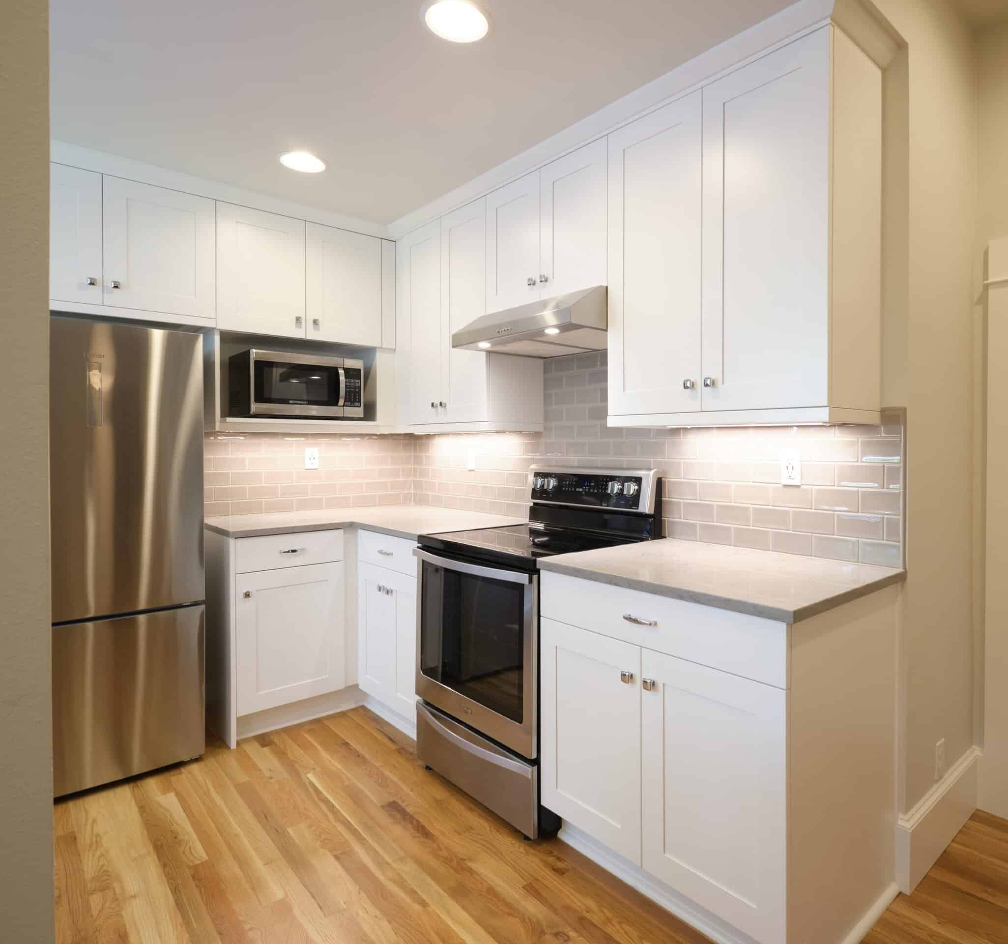 Modern kitchen with white cabinets, stainless steel appliances, light gray tile backsplash, and hardwood floor under recessed lighting.
