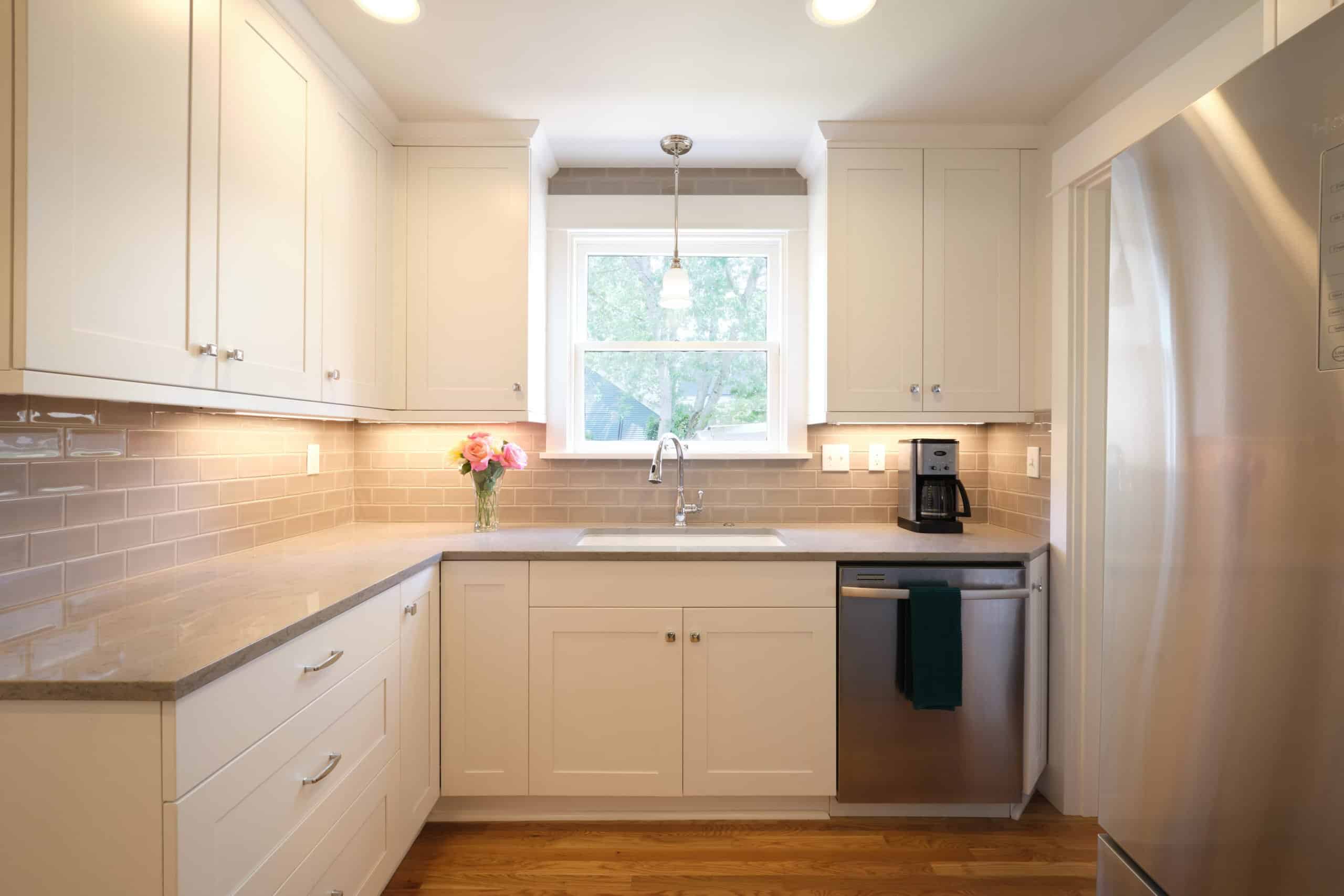 Modern kitchen with white cabinets, stainless steel appliances, a sink under a window, light tile backsplash, wood floor, and a vase of pink flowers on the countertop.