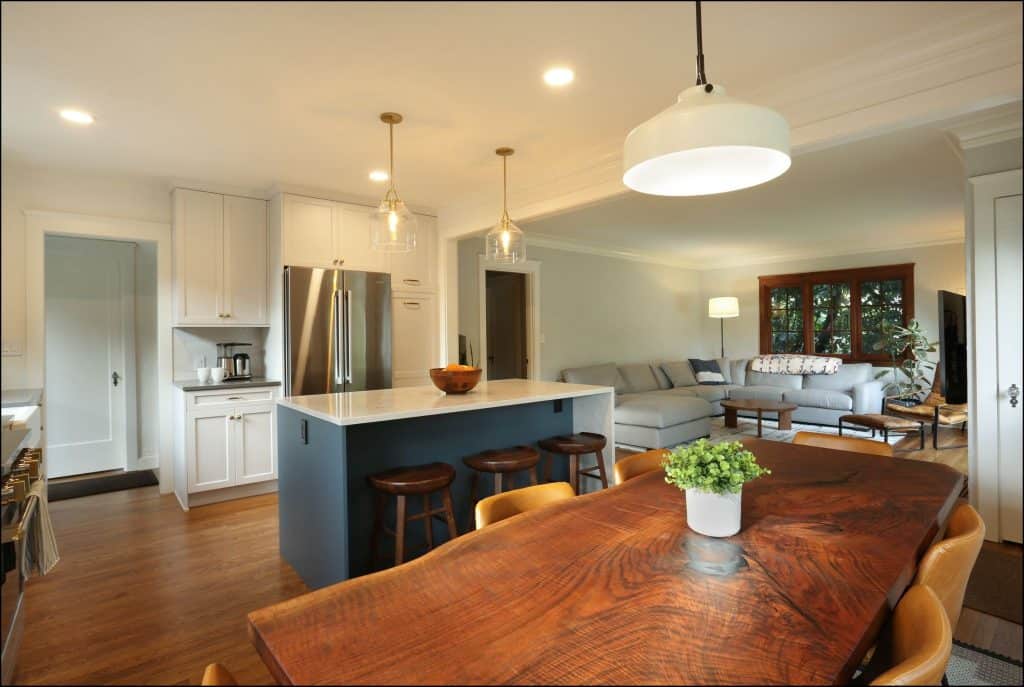 Modern open-concept kitchen and living area with a wooden dining table, island with stools, stainless steel appliances, and a sectional sofa in the background.