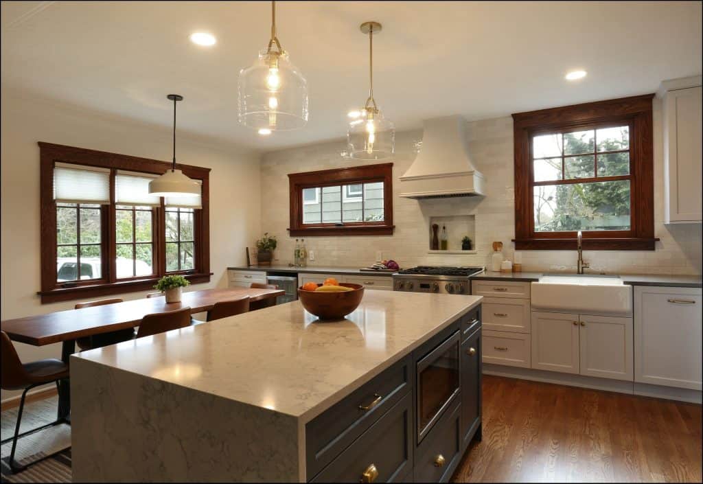 Modern kitchen with marble island, stainless steel appliances, white cabinetry, wooden floor, dining table by a window, and pendant lights overhead.
