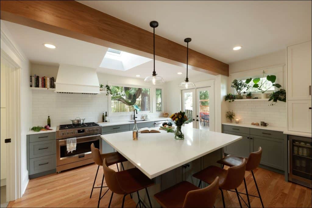 Modern kitchen with a large white island, brown leather chairs, stainless steel appliances, wooden floors, and greenery on shelves. Natural light streams in through windows and a skylight.