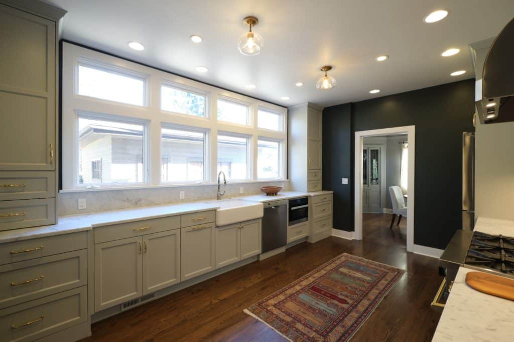 Modern kitchen with large windows, light cabinets, farmhouse sink, stainless steel appliances, marble countertops, hardwood floor, and a patterned rug. Dining room visible through doorway.