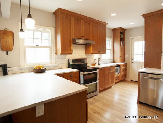 Modern kitchen with wooden cabinets, white countertops, stainless steel appliances, tile backsplash, and light wood flooring. Natural light enters through windows and a door.
